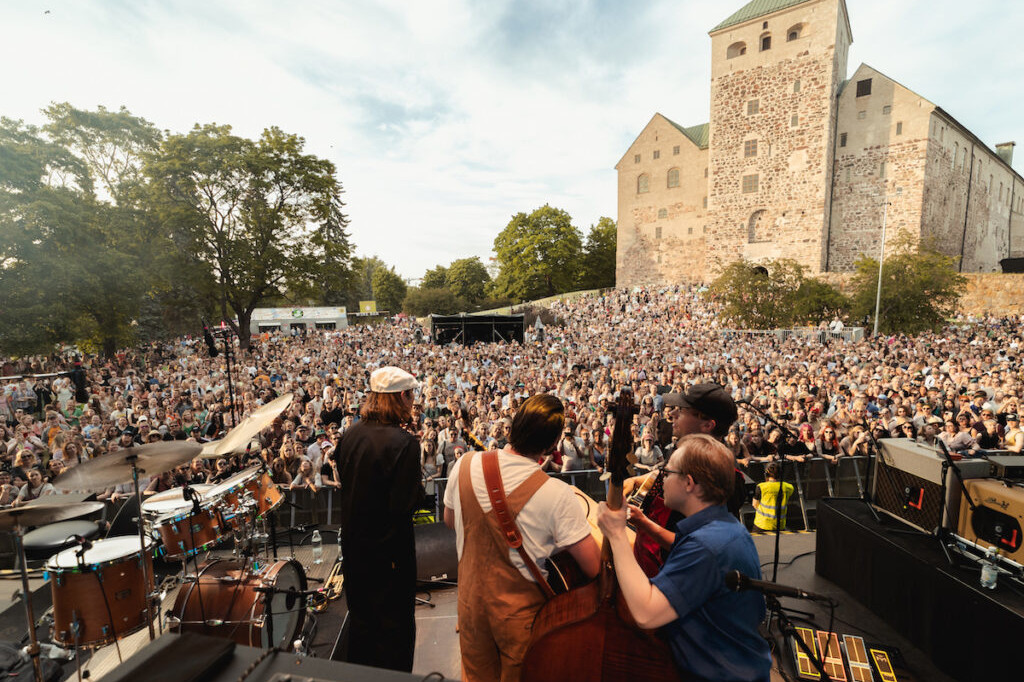 A band plays on stage in front of a large audience. An old castle is in the background.