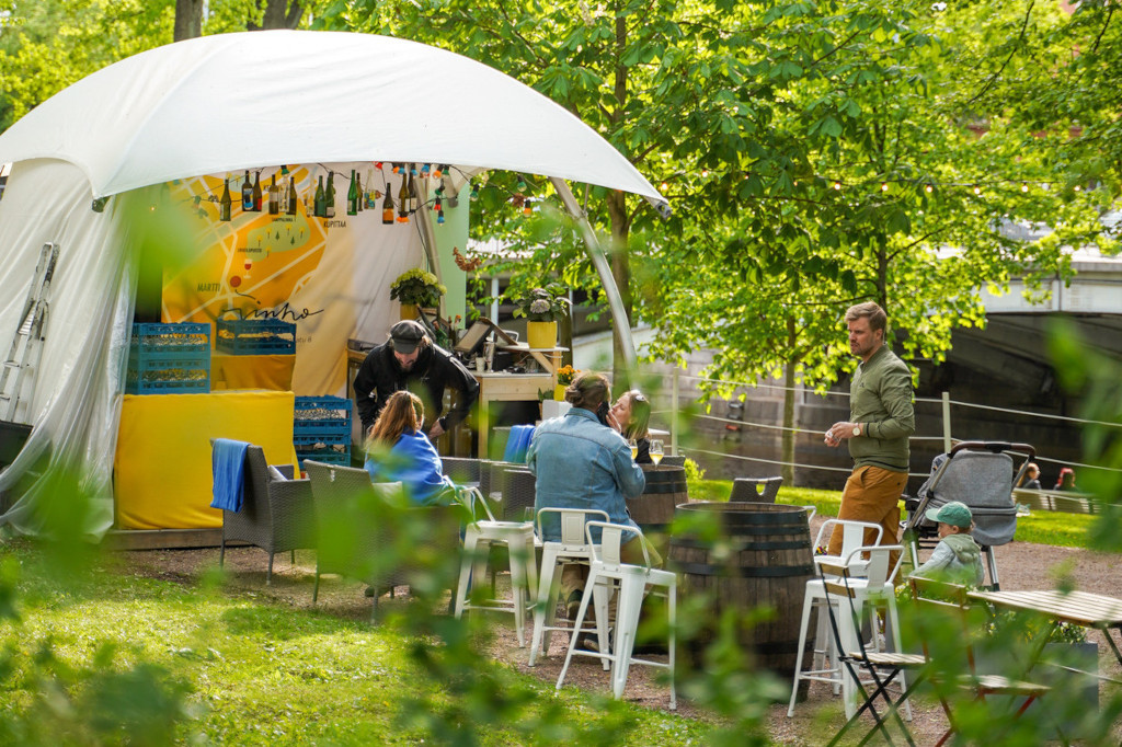 People are sitting at tables enjoying the outdoors, there is a tent and trees in the background.